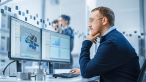 Industrial Engineer Working on a Personal Computer, Two Monitor Screens Show CAD Software with 3D Prototype of Hybrid Electric Engine and Charts. Working Modern Factory.